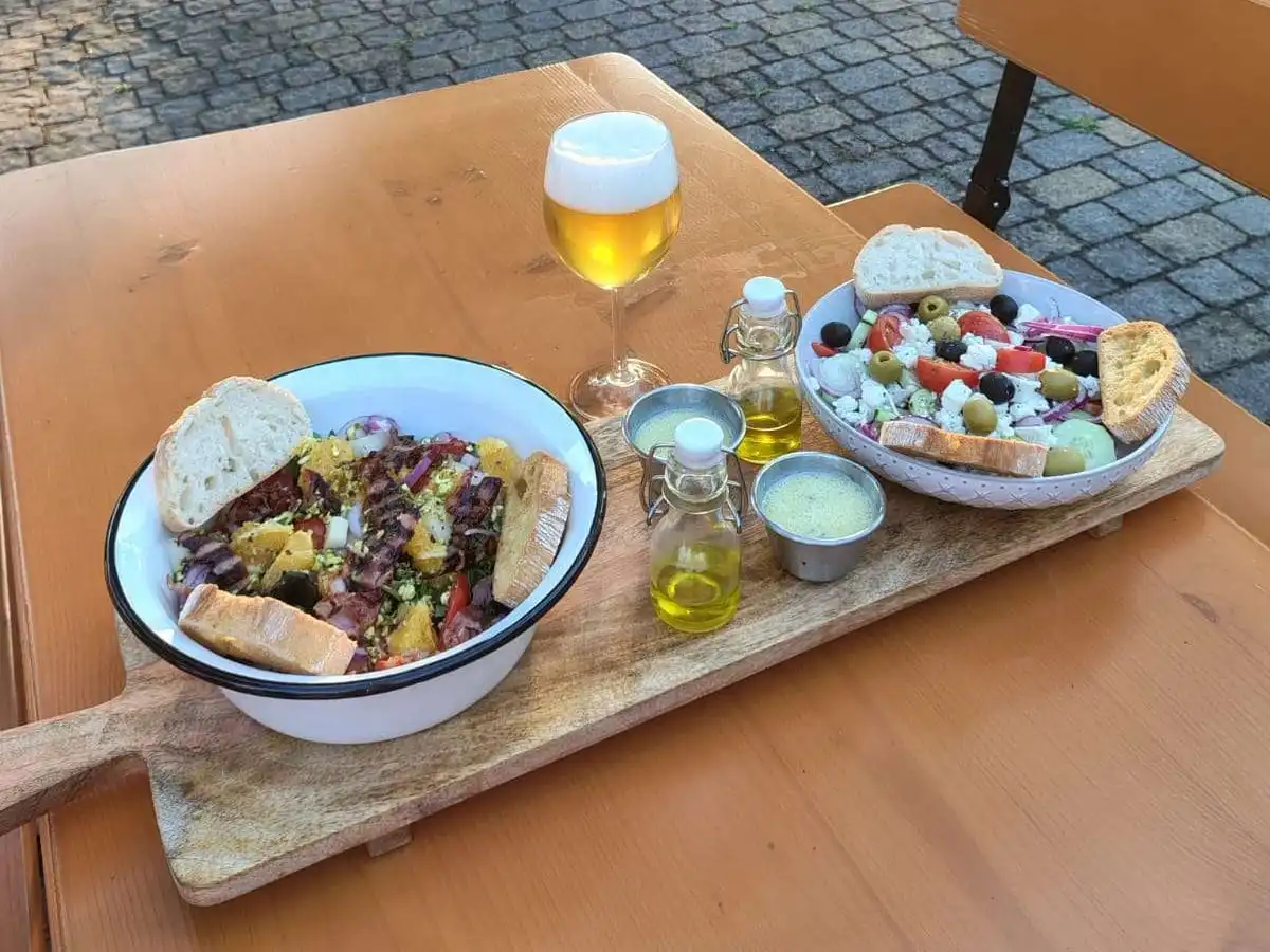 Un tagliere su un tavolo di legno in terrazza. Sopra ci sono due insalate con il pane, l'olio e un bicchiere di birra.