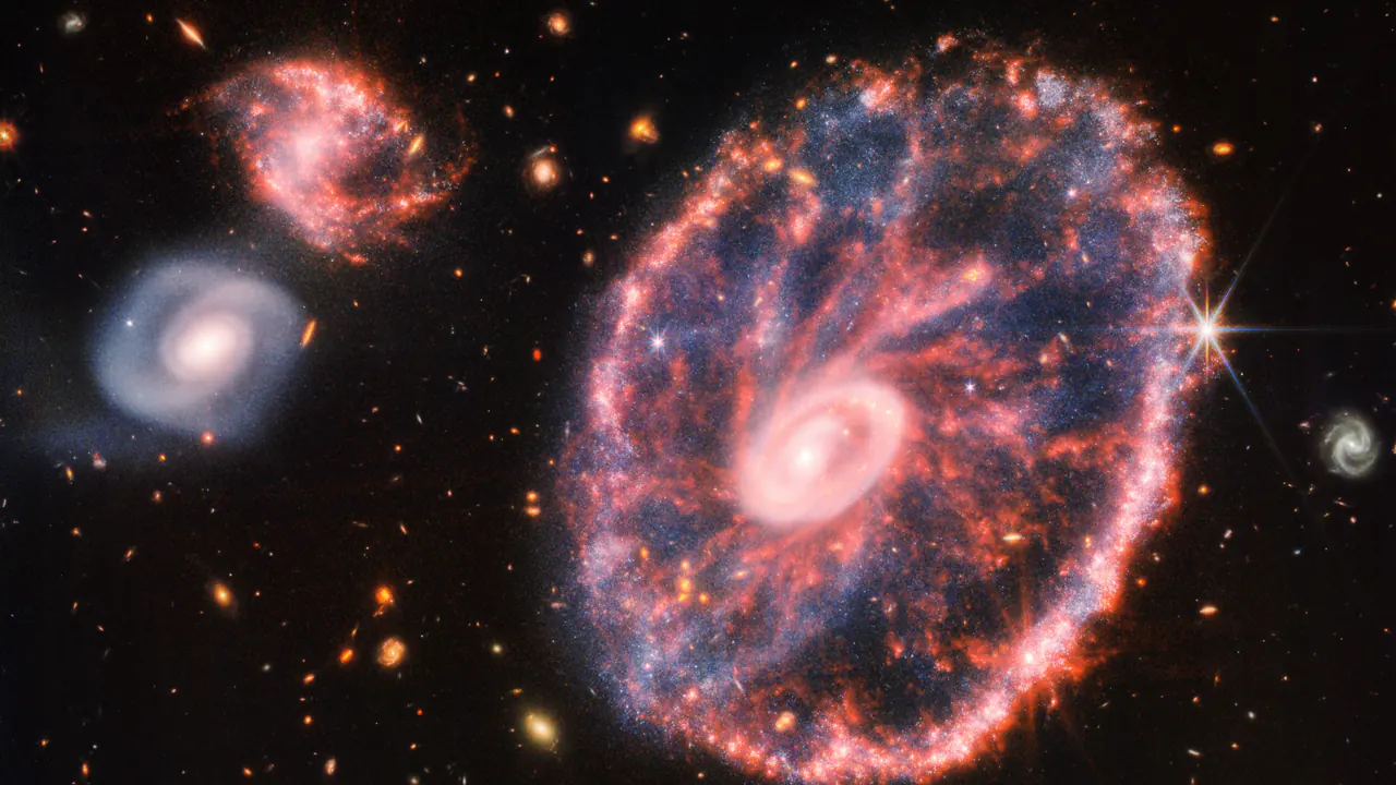 Una grande galassia ad anello, rosa e blu, con un nucleo luminoso al centro e raggi sottili. Accanto ci sono due galassie più piccole, su un fondo nero.