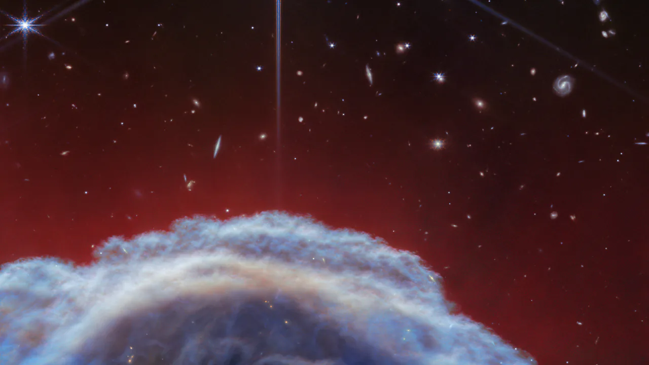 Il profilo chiaro e sfrangiato di una nube di gas e polvere che sale dal basso su un fondo rosso scuro e nero. Sopra ci sono una stella luminosa e molte galassie lontane: è la cima della Nebulosa Testa di Cavallo.
