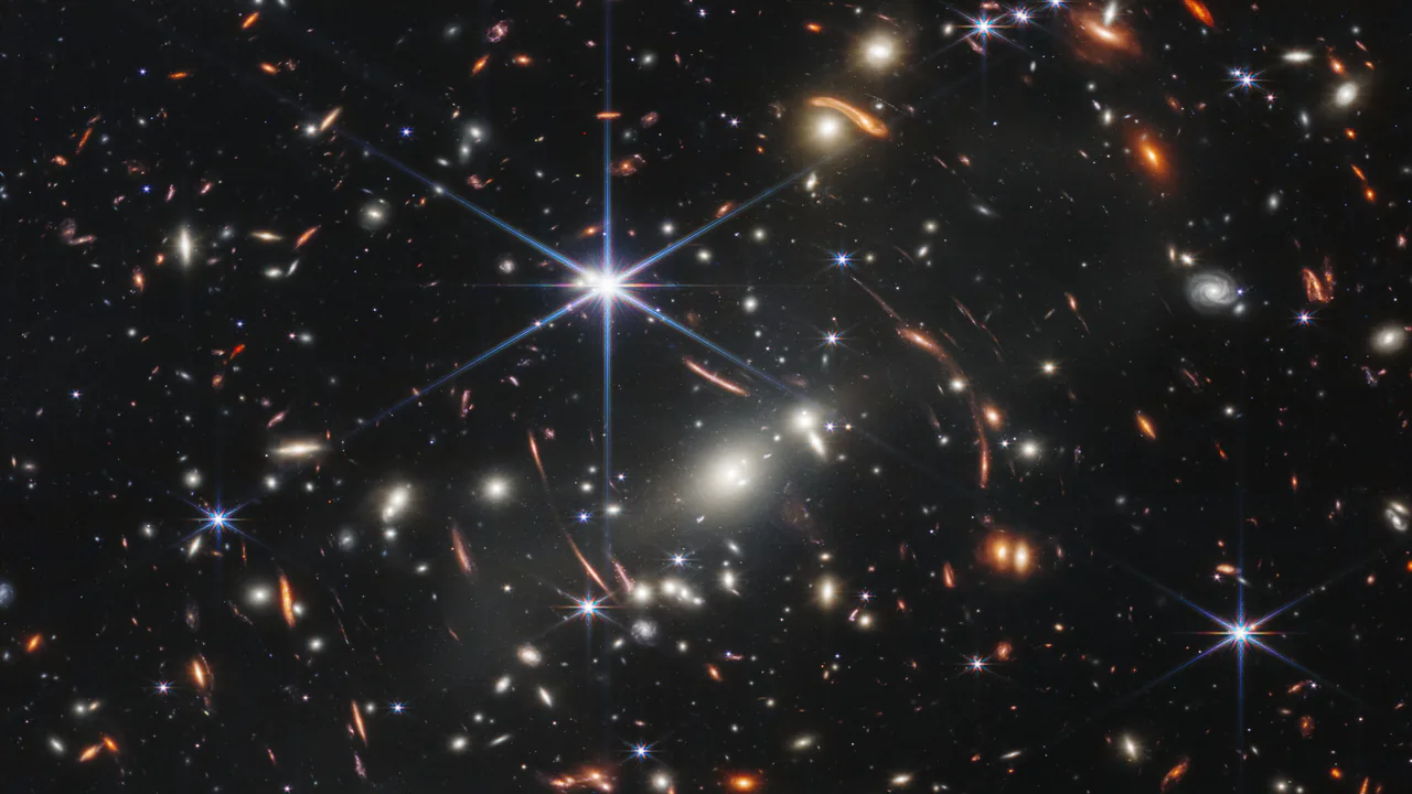 Un campo nero fitto di galassie lontanissime, bianche, arancioni e rosse. Alcune sono piegate in archi sottili attorno a un ammasso luminoso al centro.