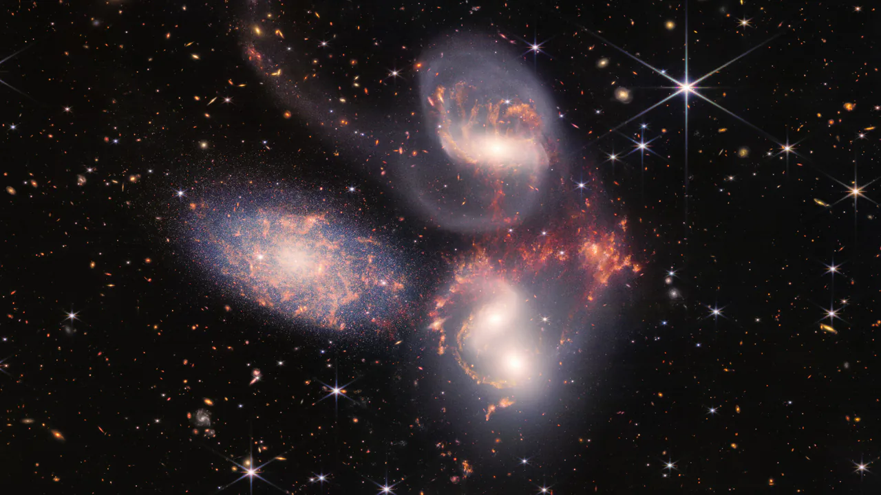 Un gruppo di galassie su un fondo nero pieno di galassie minute. Alcune si sfiorano e si deformano a vicenda, con scie di gas rosso e bianco.