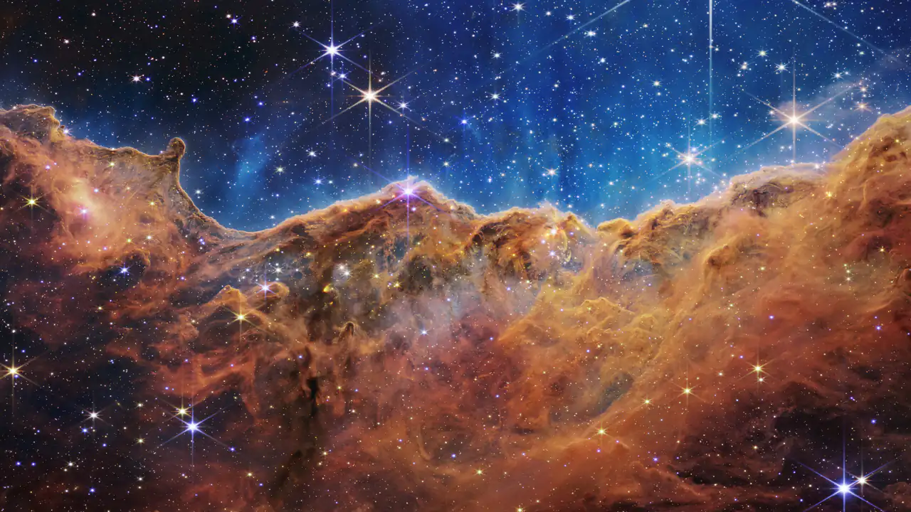 Un crinale di gas e polvere color rame, punteggiato di stelle luminose, sotto un cielo blu scuro pieno di stelle. È il bordo di una regione in cui nascono stelle, nella Nebulosa della Carena.